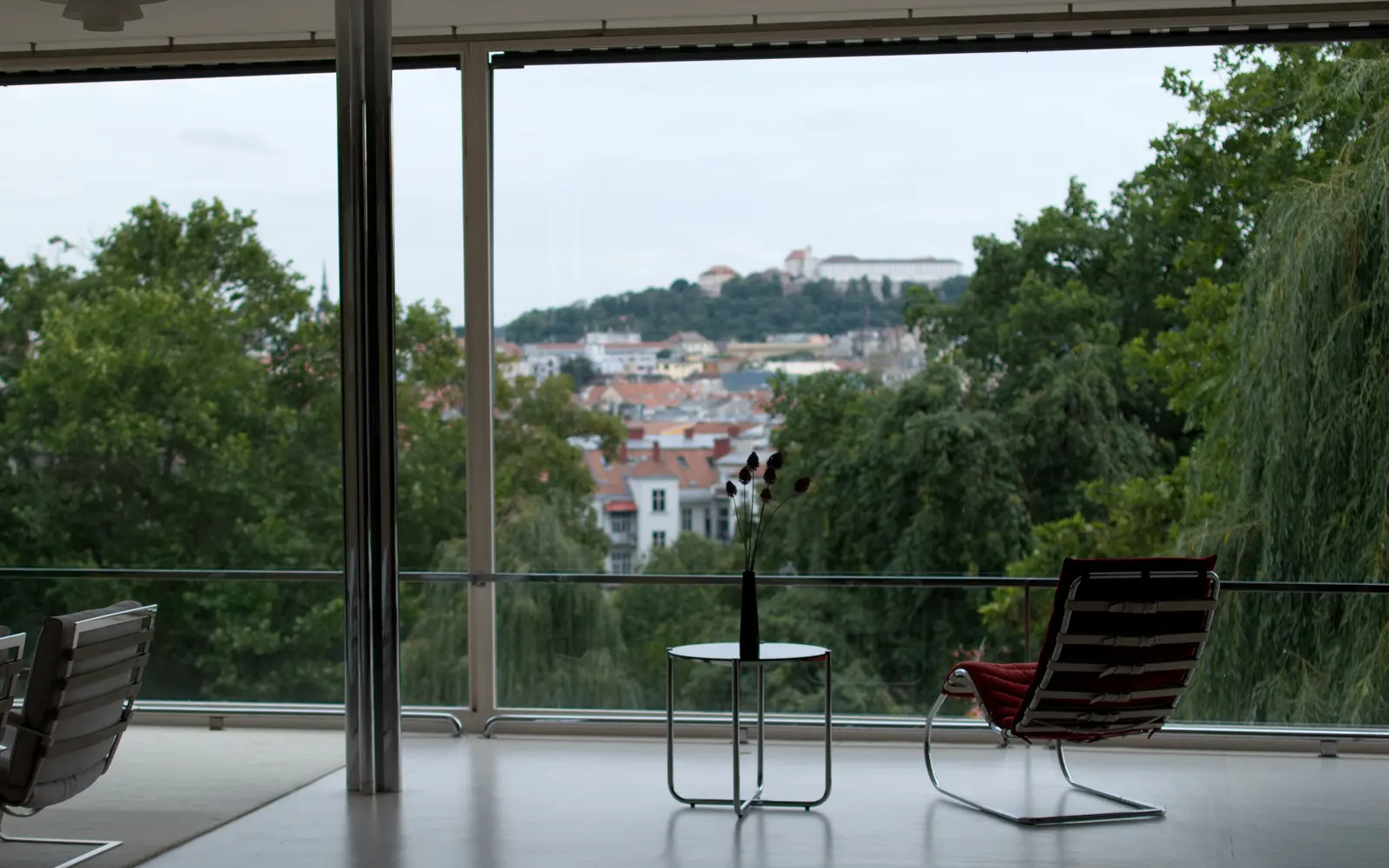 The living-room glass wall, a chrome column and a red lounge chair, with trees and Špilberk Castle beyond